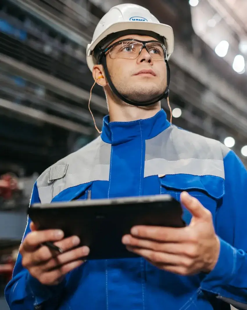 Supervisor de planta con casco, gafas de seguridad y tapones auditivos anotando en una tableta dentro de una nave industrial — contexto de reclutamiento industrial de Indu Talent.